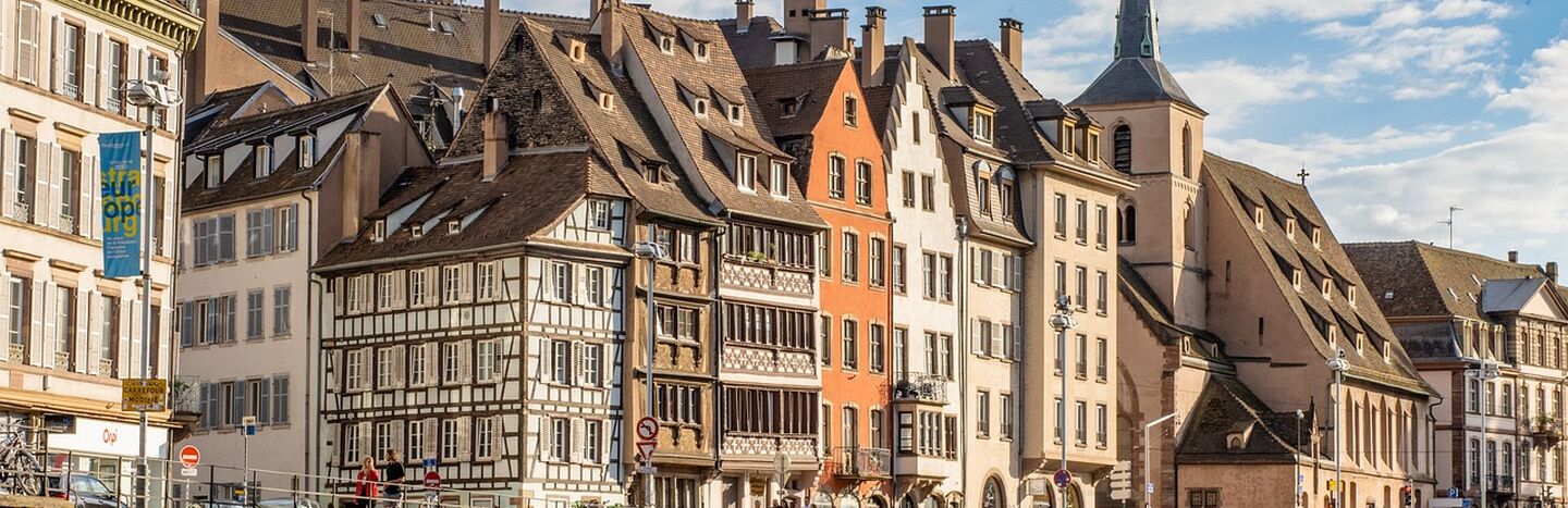 Bunte historische Gebäude und eine Kirche säumen einen Kanal unter einem teilweise bewölkten Himmel in einer europäischen Stadt.