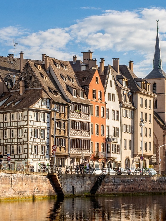 Bunte historische Gebäude und eine Kirche säumen einen Kanal unter einem teilweise bewölkten Himmel in einer europäischen Stadt.