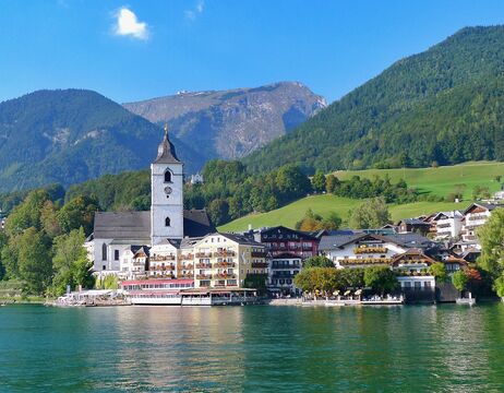 Ein Dorf am See mit einem Kirchturm, bunten Gebäuden und Bergen im Hintergrund.