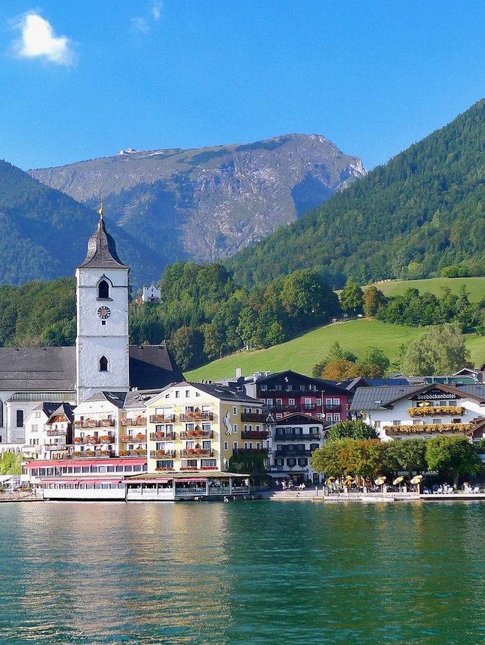Ein Dorf am See mit einem Kirchturm, bunten Gebäuden und Bergen im Hintergrund.