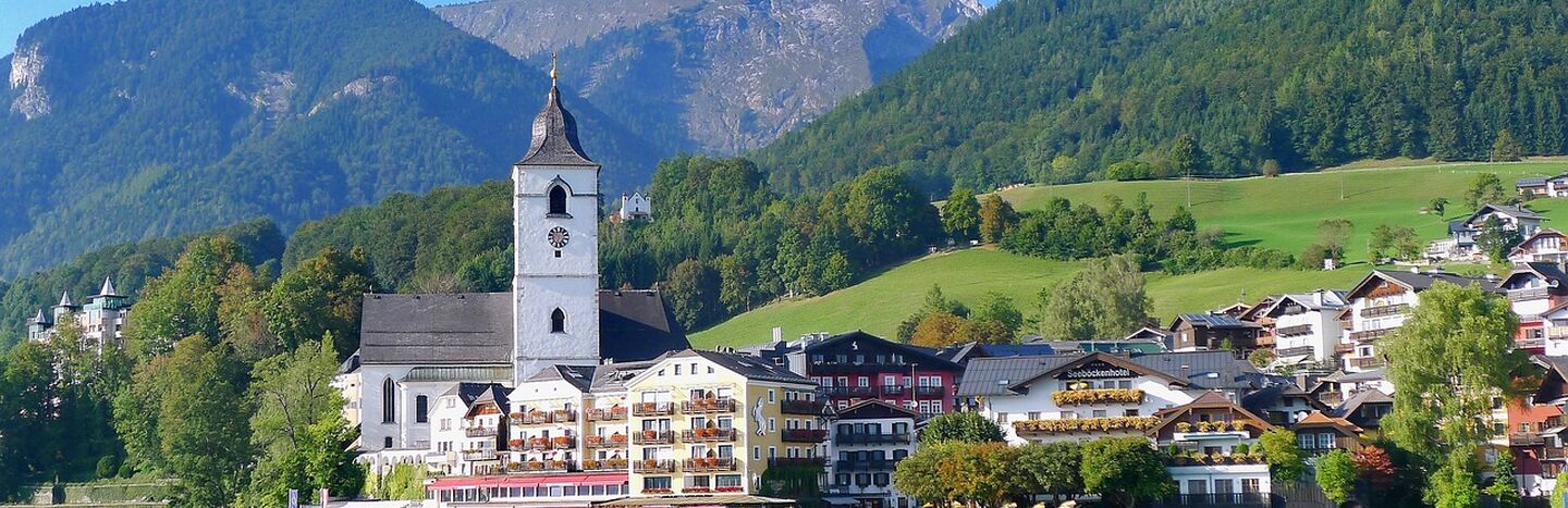 Ein Dorf am See mit einem Kirchturm, bunten Gebäuden und Bergen im Hintergrund.