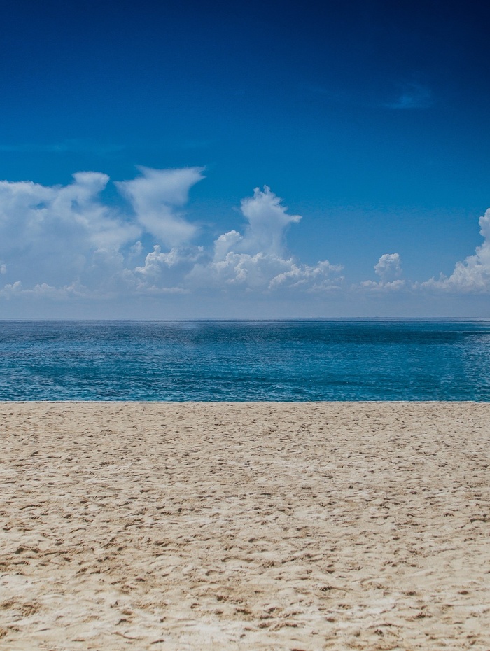 Sandstrand mit ruhigem blauen Meer unter einem klaren Himmel mit vereinzelten Wolken.