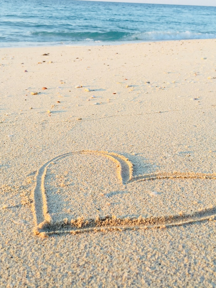 Eine Person mit Hut sitzt auf einer felsigen Klippe und blickt unter einem blauen Himmel mit Wolkenfetzen auf den Ozean hinaus.