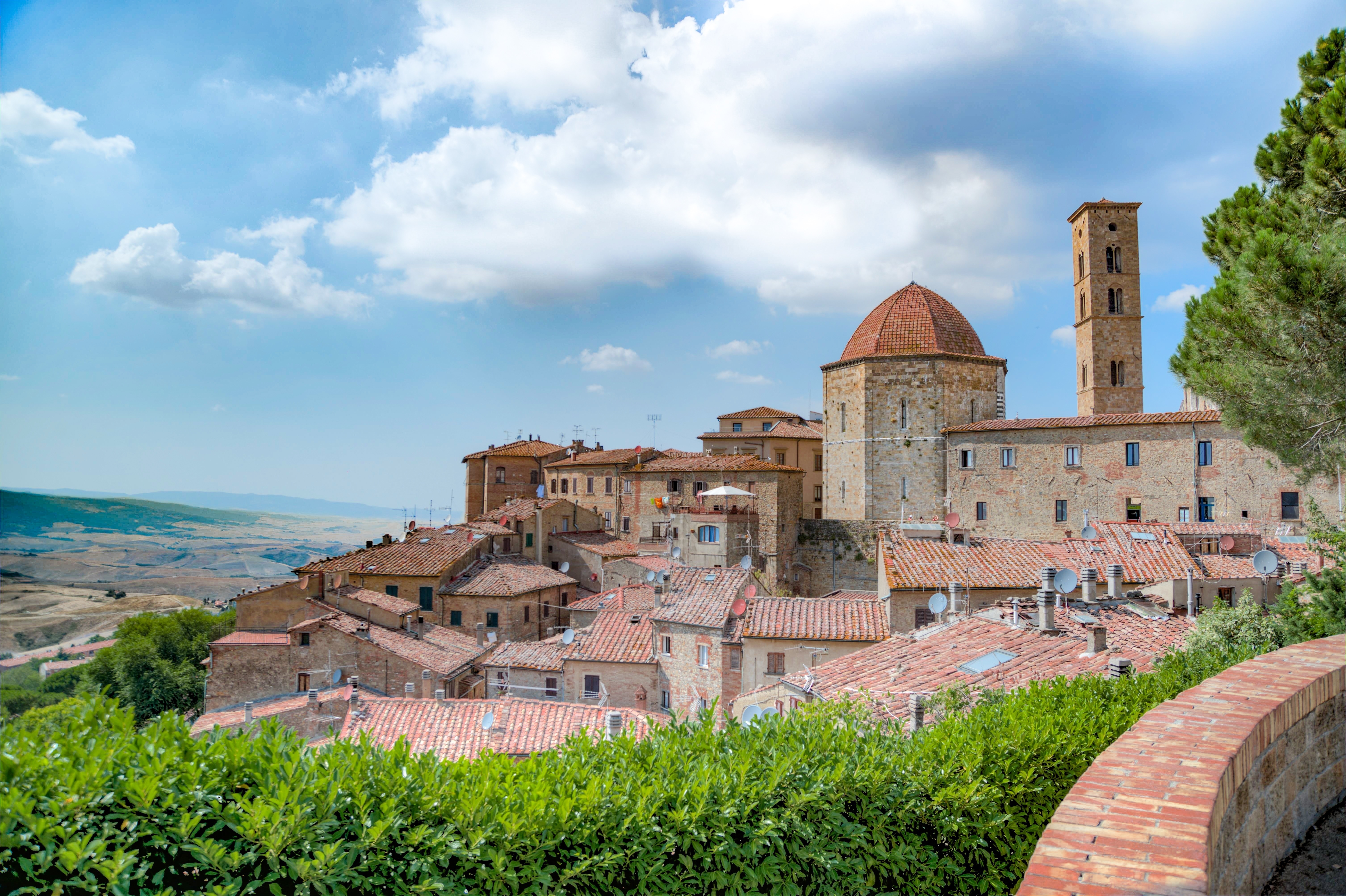 Italienisches Dorf auf einem Hügel mit Terrakotta-Dächern, Steinhäusern und einer Kirche unter einem blauen Wolkenhimmel.