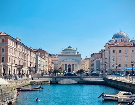Kanal mit kleinen Booten, pastellfarbenen Gebäuden und einer Kuppelkirche unter einem klaren blauen Himmel in einer italienischen Stadt.