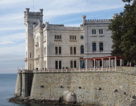 Die weiße Steinburg mit ihren Türmen steht auf einer Klippe am Meer, teilweise im Schatten von Bäumen, unter einem blauen Himmel.