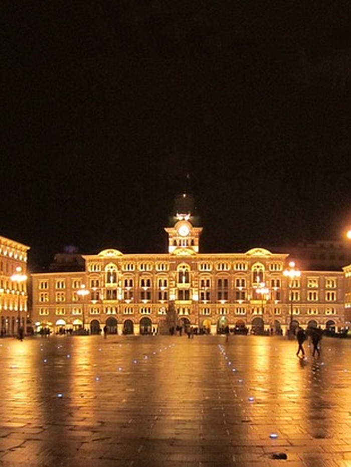 Ein großes beleuchtetes Gebäude auf einem nächtlichen Stadtplatz mit spazierenden Menschen und leuchtenden Straßenlaternen.