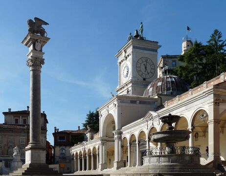 Historischer Platz mit Uhrenturm, statuengekrönter Säule, Bögen und Springbrunnen unter strahlend blauem Himmel.