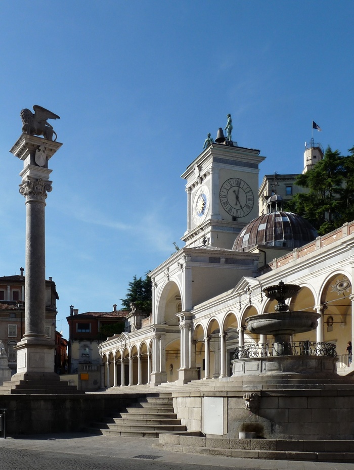 Historischer Platz mit Uhrenturm, statuengekrönter Säule, Bögen und Springbrunnen unter strahlend blauem Himmel.