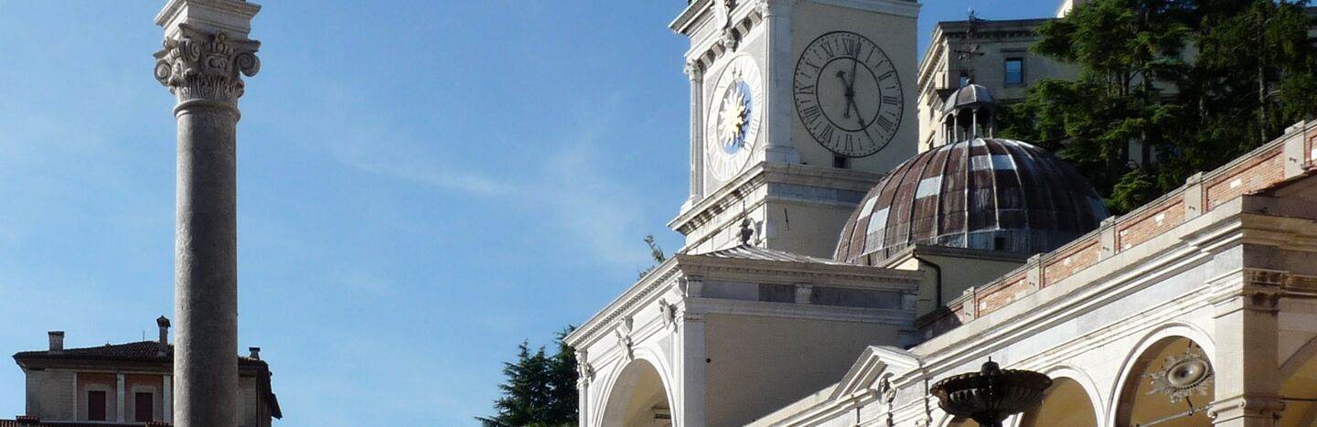 Historischer Platz mit Uhrenturm, statuengekrönter Säule, Bögen und Springbrunnen unter strahlend blauem Himmel.