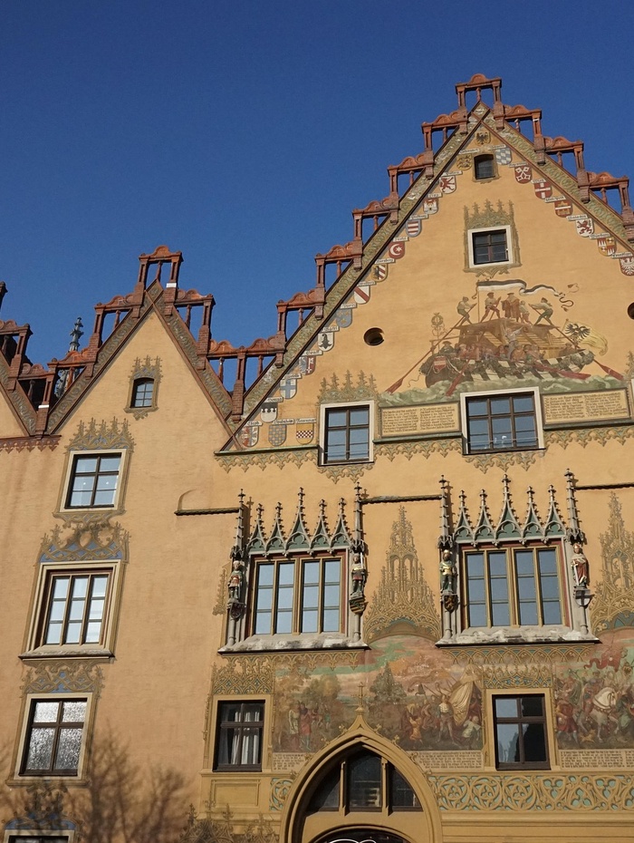 Verziertes Gebäude mit Wandmalerei, detaillierten Fenstern und steilem Giebeldach vor einem klaren blauen Himmel.