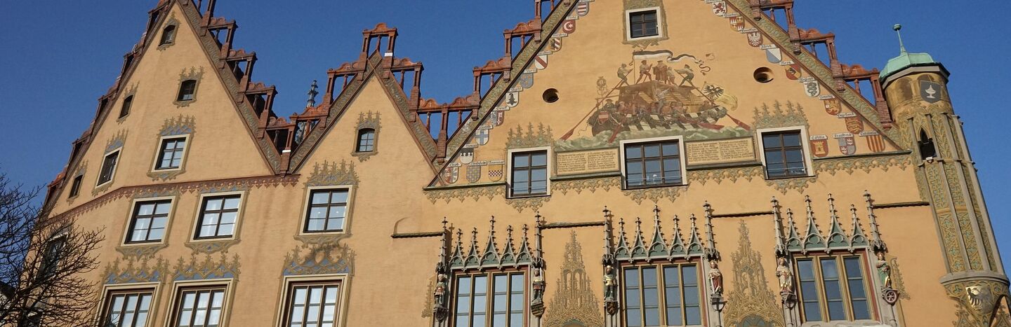 Verziertes Gebäude mit Wandmalerei, detaillierten Fenstern und steilem Giebeldach vor einem klaren blauen Himmel.