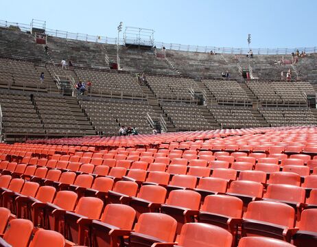 Reihen leerer roter Sitze im Vordergrund eines großen, meist leeren Freiluftstadions unter klarem Himmel.