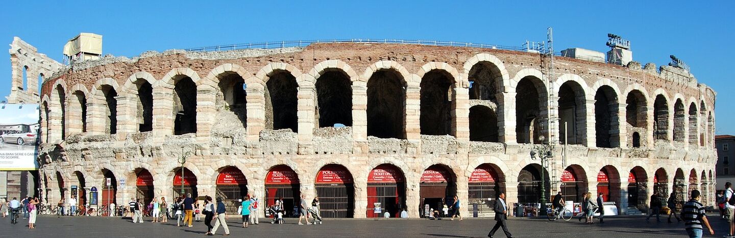 Antikes römisches Amphitheater mit Steinbögen und Menschen, die an einem sonnigen Tag davor spazieren gehen.