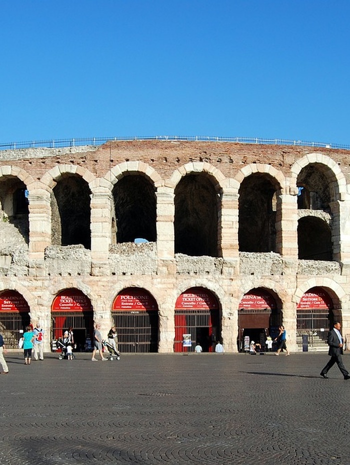 Antikes römisches Amphitheater mit Steinbögen und Menschen, die an einem sonnigen Tag davor spazieren gehen.