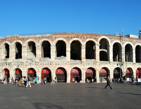 Antikes römisches Amphitheater mit Steinbögen und Menschen, die an einem sonnigen Tag davor spazieren gehen.
