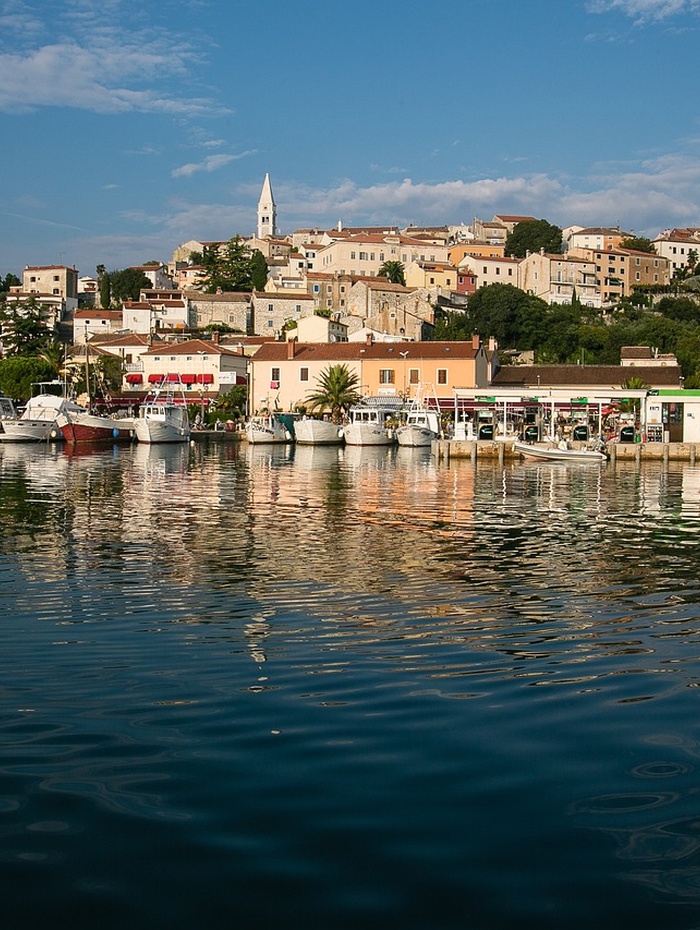 Bunte Häuser und Boote am Wasser, mit einem Dorf auf einem Hügel und einem Kirchturm im Hintergrund.