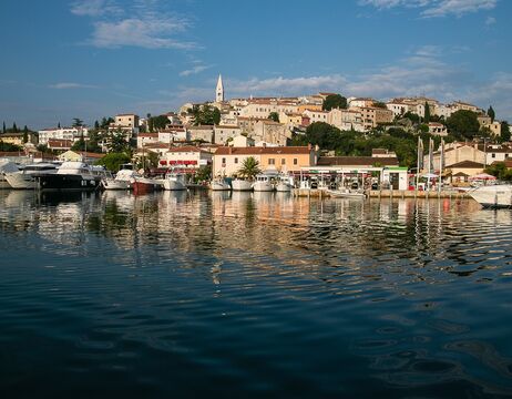 Bunte Häuser und Boote am Wasser, mit einem Dorf auf einem Hügel und einem Kirchturm im Hintergrund.