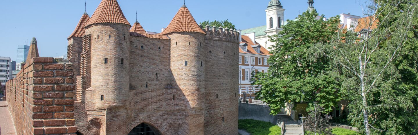 Ein historisches Stadttor aus Backstein mit roten Dächern, umgeben von Grünflächen und nahe gelegenen Kirchtürmen unter einem klaren Himmel.