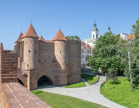 Ein historisches Stadttor aus Backstein mit roten Dächern, umgeben von Grünflächen und nahe gelegenen Kirchtürmen unter einem klaren Himmel.