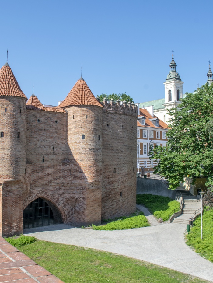 Ein historisches Stadttor aus Backstein mit roten Dächern, umgeben von Grünflächen und nahe gelegenen Kirchtürmen unter einem klaren Himmel.