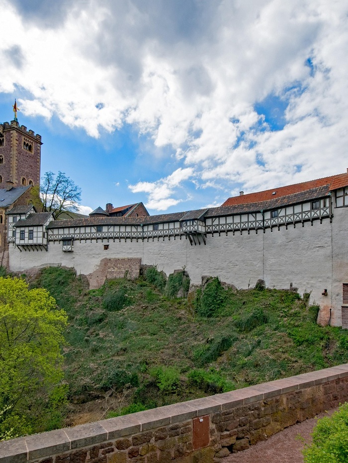 Eine Burgmauer aus Stein und Holz mit einem Turm, unter einem teilweise bewölkten blauen Himmel.