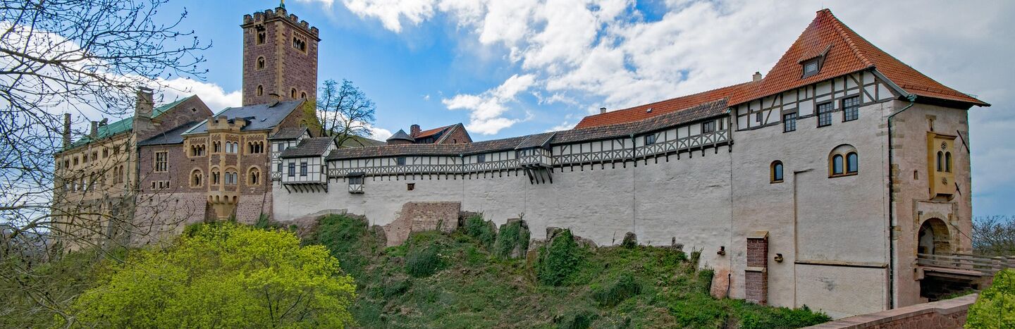 Eine Burgmauer aus Stein und Holz mit einem Turm, unter einem teilweise bewölkten blauen Himmel.