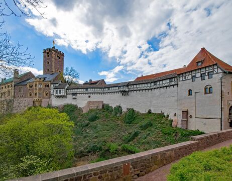 Eine Burgmauer aus Stein und Holz mit einem Turm, unter einem teilweise bewölkten blauen Himmel.