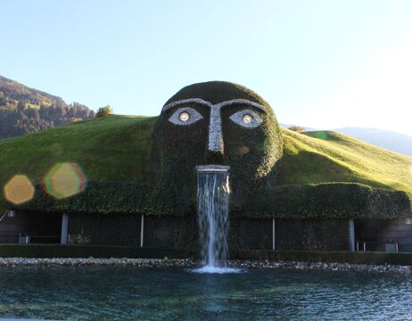 Eine Hangskulptur eines Gesichts, aus dessen Mund Wasser in ein Becken fließt, umgeben von Grünpflanzen.