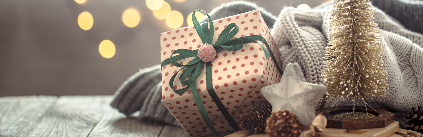 Polka-Dot-Geschenkbox mit grünem Band, kuscheliger Decke, Stern-Ornament und warmen Lichterketten im Hintergrund.
