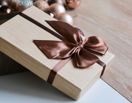 Geschenkkarton mit braunem Band, daneben Ornamente, ein kleiner Baum und eine brennende Kerze auf einem Holztisch.
