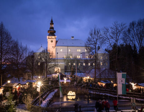Ein großes, schneebedecktes Schloss, das in der Abenddämmerung mit festlichen Lichtern beleuchtet ist und unter dem sich Menschen versammelt haben.