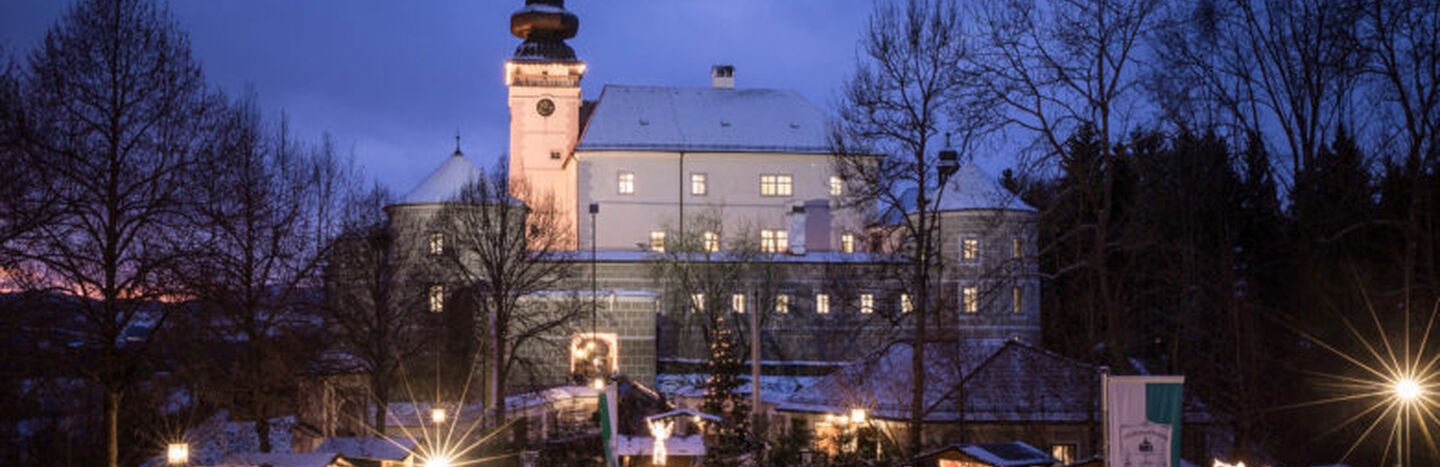 Ein großes, schneebedecktes Schloss, das in der Abenddämmerung mit festlichen Lichtern beleuchtet ist und unter dem sich Menschen versammelt haben.