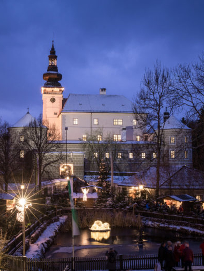 Ein großes, schneebedecktes Schloss, das in der Abenddämmerung mit festlichen Lichtern beleuchtet ist und unter dem sich Menschen versammelt haben.