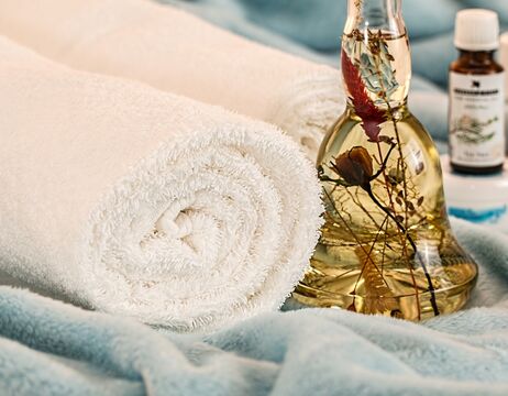 Aufgerolltes weißes Handtuch neben einer Glasflasche mit getrockneten Blumen, die auf einem weichen blauen Stoff liegt.
