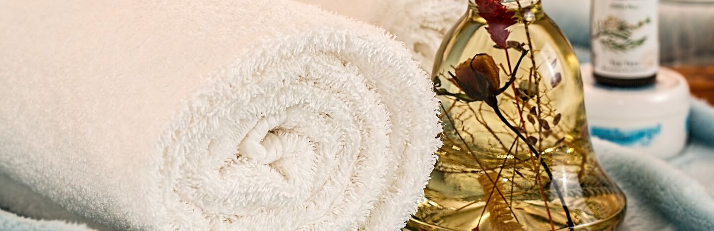 Aufgerolltes weißes Handtuch neben einer Glasflasche mit getrockneten Blumen, die auf einem weichen blauen Stoff liegt.