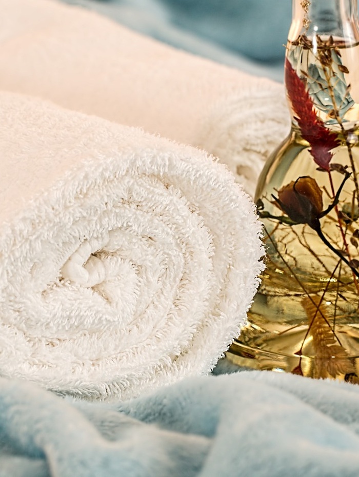 Aufgerolltes weißes Handtuch neben einer Glasflasche mit getrockneten Blumen, die auf einem weichen blauen Stoff liegt.