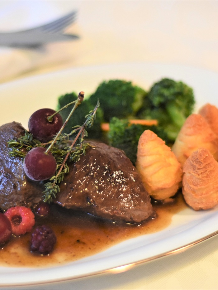 Ein Tellergericht mit Rindfleisch, Soße, Beeren, Brokkoli und drei Herzoginkartoffeln auf einem weißen Teller.