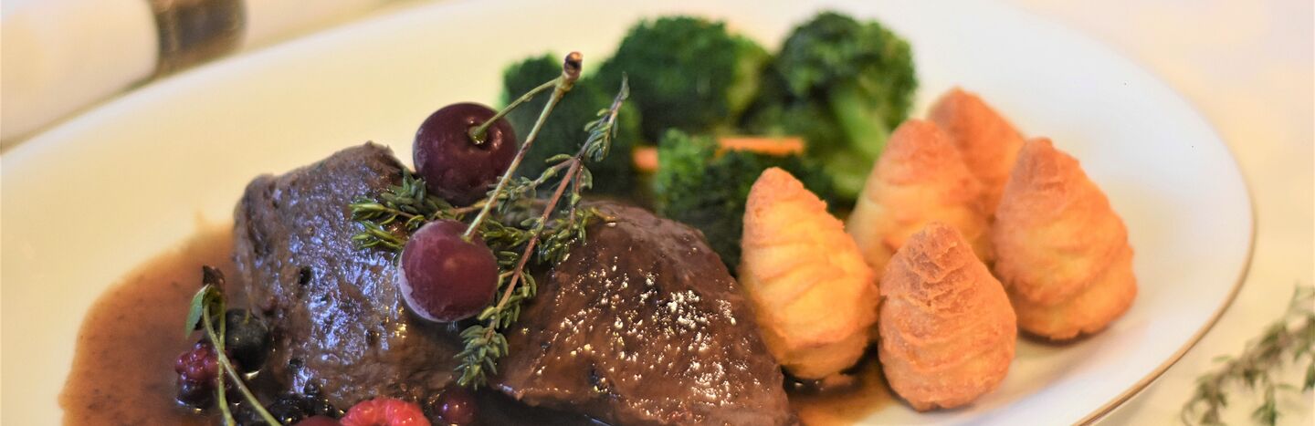 Ein Tellergericht mit Rindfleisch, Soße, Beeren, Brokkoli und drei Herzoginkartoffeln auf einem weißen Teller.