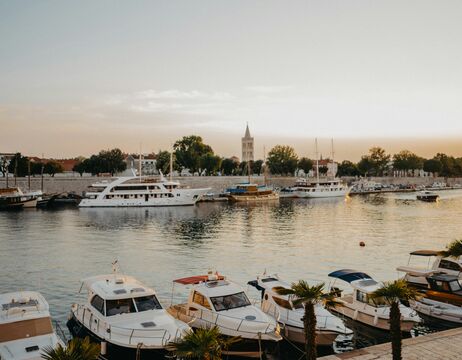 Boote und Yachten in einem ruhigen Hafen bei Sonnenuntergang mit Bäumen und einem Kirchturm im Hintergrund.