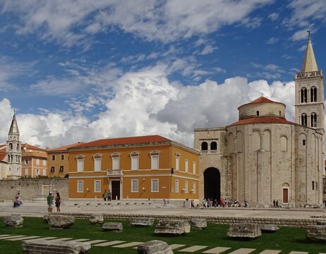 Alte Steinruinen mit historischen Gebäuden und einem Kirchturm unter einem teilweise bewölkten blauen Himmel.