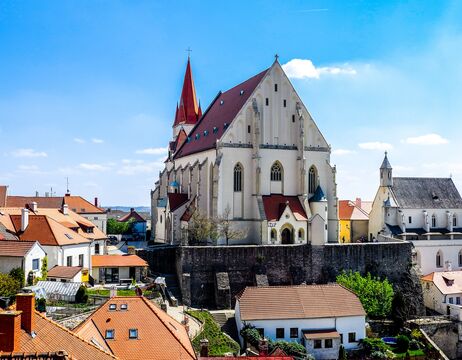 Gotische Kirche mit rotem Dach und hohem Kirchturm, umgeben von Häusern unter blauem Himmel.