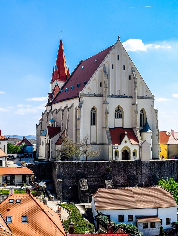 Gotische Kirche mit rotem Dach und hohem Kirchturm, umgeben von Häusern unter blauem Himmel.