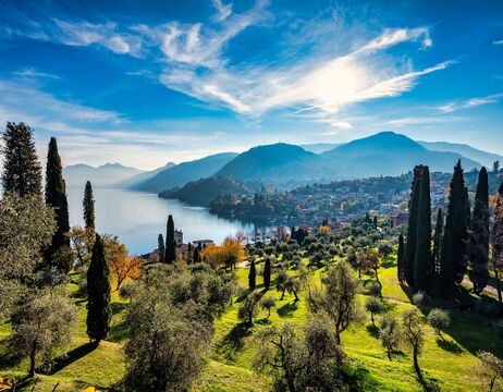 Sonnige Landschaft mit einem See, grünen Hügeln, hohen Bäumen und fernen Bergen unter blauem Himmel.