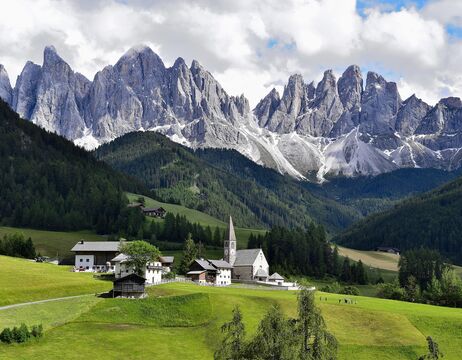 Ein kleines Dorf mit einer Kirche liegt in einem grünen Tal, das von steilen, zerklüfteten grauen Bergen unter einem bewölkten Himmel umgeben ist.