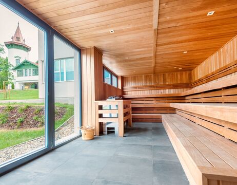 Moderner Saunaraum aus Holz mit großen Fenstern und Blick auf eine Graslandschaft und ein Gebäude mit einem Turm im Freien.