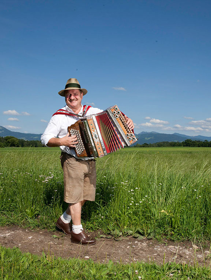 Ein Mann in traditioneller bayerischer Kleidung spielt ein Akkordeon auf einer Wiese mit Bergen im Hintergrund.