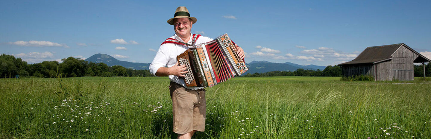 Ein Mann in traditioneller bayerischer Kleidung spielt ein Akkordeon auf einer Wiese mit Bergen im Hintergrund.