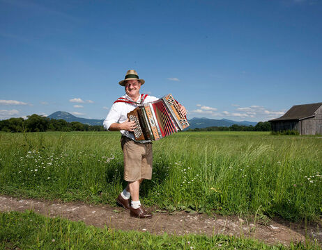Ein Mann in traditioneller bayerischer Kleidung spielt ein Akkordeon auf einer Wiese mit Bergen im Hintergrund.