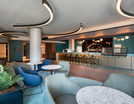 Moderne Lounge mit gebogener Beleuchtung, runden Tischen, Sesseln und einer Bar mit hohen Hockern im Hintergrund.